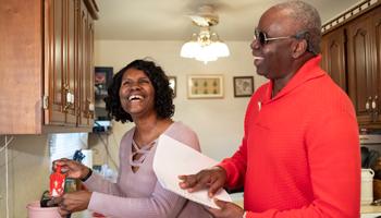 A husband a wife smile together in the kitchen. The man reads a recipe to his wife from a Braille page. 